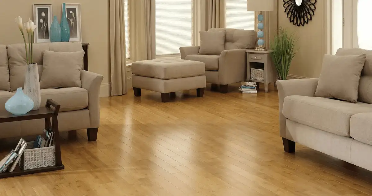 A living room with professionally installed bamboo flooring, beige sofas, a matching ottoman, a side table with a blue lamp, vases, framed pictures, and large windows with blinds and curtains in Akron, OH